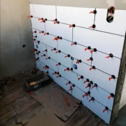 Układanie białych płytek na ścianie z użyciem systemów poziomowania, widoczne gniazdko elektryczne i drewnopodobna podłoga.