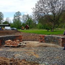 Ricobut - Budowa muru z kamienia i czerwonej cegły w Gryfowie Śląskim, widoczny pracownik w trakcie rozmowy telefonicznej, na palecie ułożone cegły, w tle zieleń i zaparkowane pojazdy.