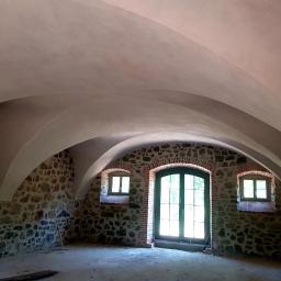 Ricobut - Wnętrze pomieszczenia z kamiennymi ścianami i łukowym sufitem po szpachlowaniu, widoczne okna i drzwi z drewnianą stolarką.