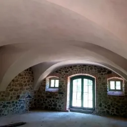 Wnętrze pomieszczenia z kamiennymi ścianami i łukowym sufitem po szpachlowaniu, widoczne okna i drzwi z drewnianą stolarką.