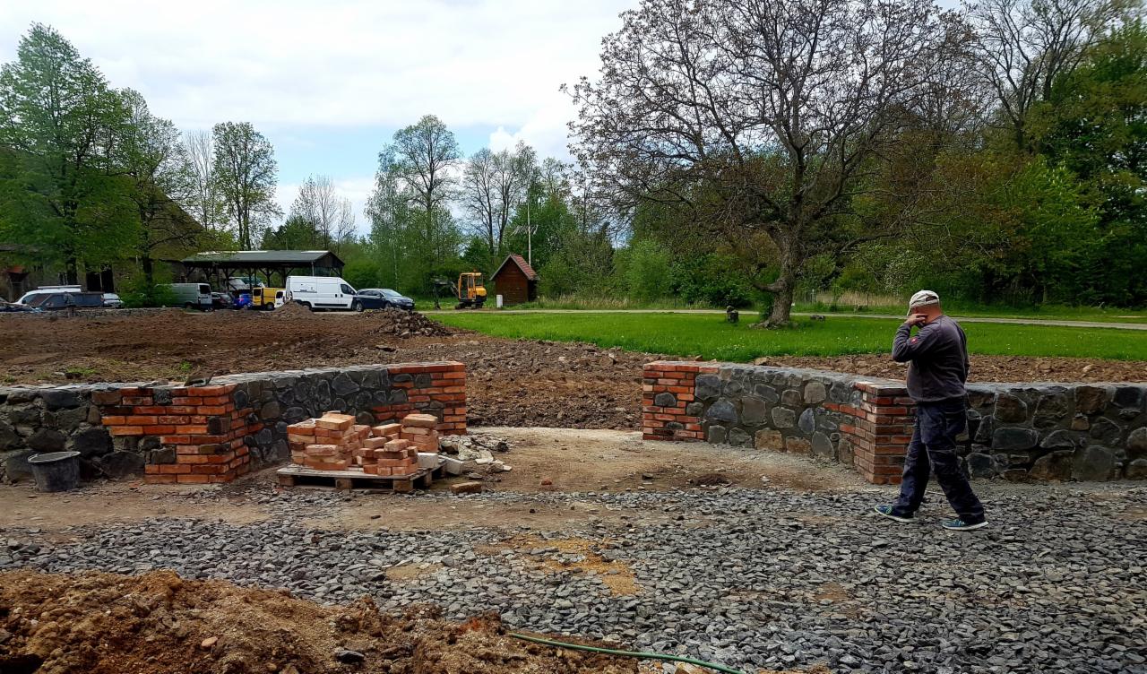 Budowa muru z kamienia i czerwonej cegły w Gryfowie Śląskim, widoczny pracownik w trakcie rozmowy telefonicznej, na palecie ułożone cegły, w tle zieleń i zaparkowane pojazdy.