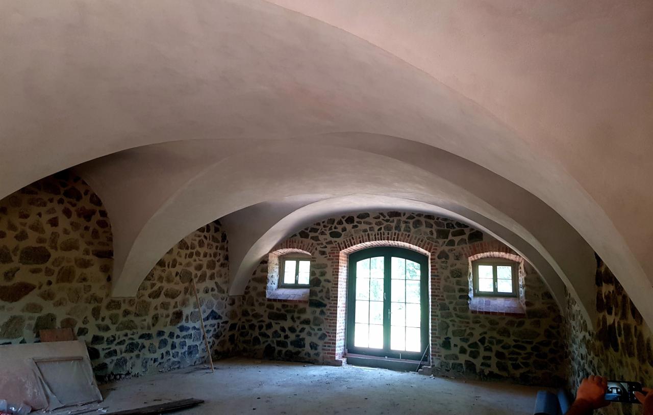 Wnętrze pomieszczenia z kamiennymi ścianami i łukowym sufitem po szpachlowaniu, widoczne okna i drzwi z drewnianą stolarką.