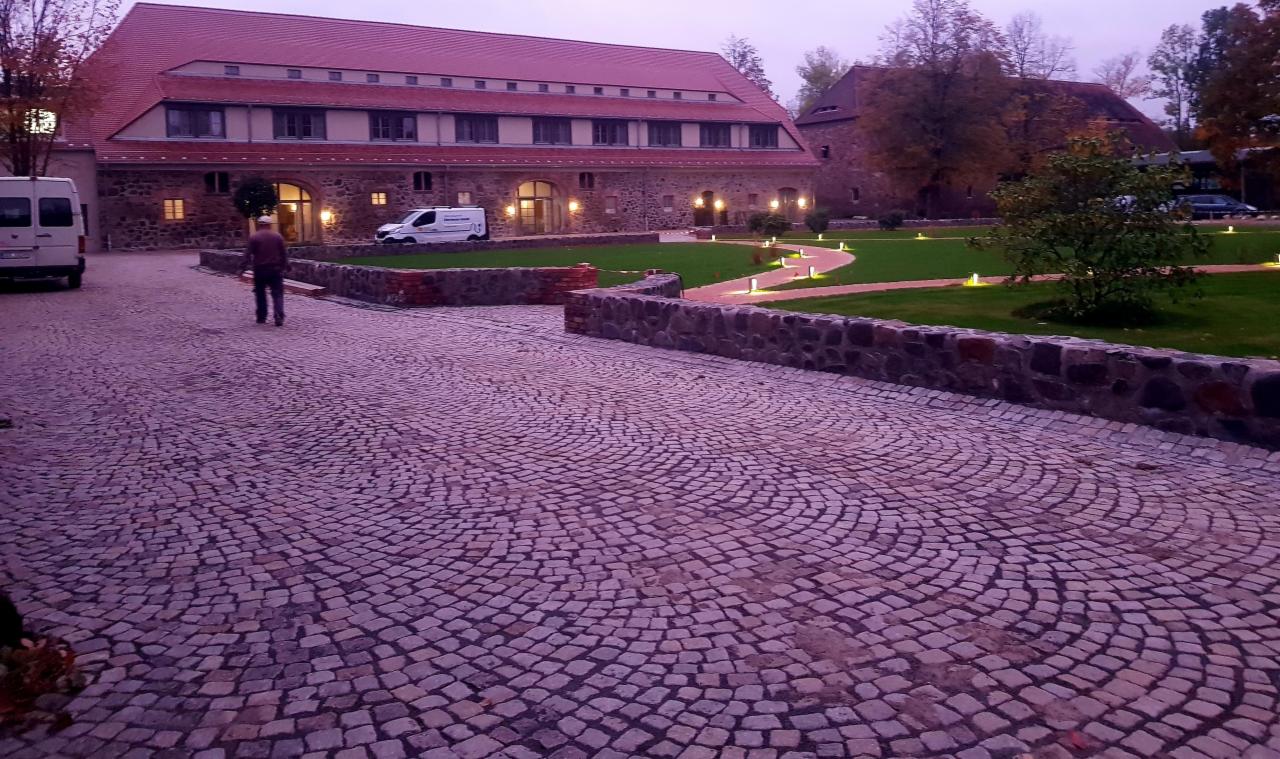 Dziedziniec z brukowaną nawierzchnią ułożoną w koncentryczne łuki, oświetlony małymi lampkami, z budynkiem z czerwonym dachem w tle i osobą idącą w jego kierunku.