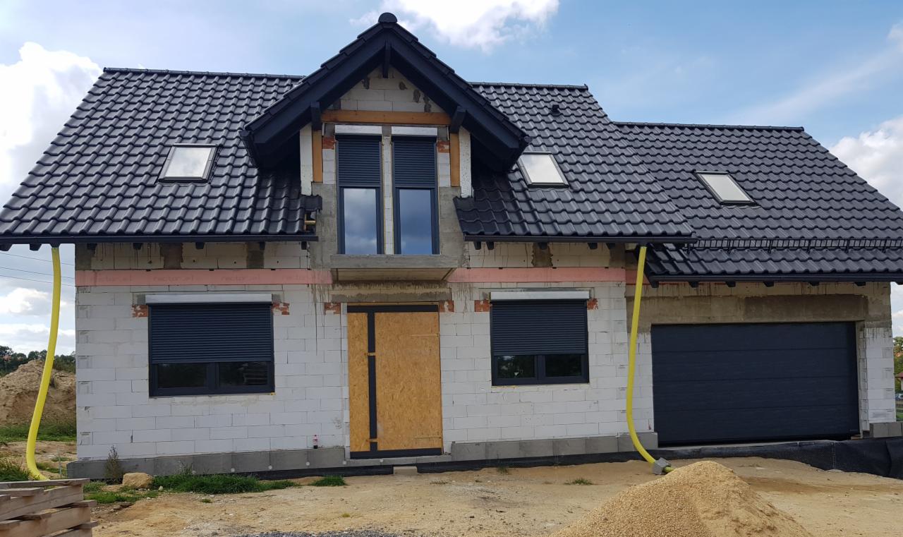 Surowy stan domu z białego bloczka, czarną dachówką i roletami zewnętrznymi, z widoczną deską zamiast drzwi wejściowych i żółtym wężem wentylacyjnym wzdłuż ściany.