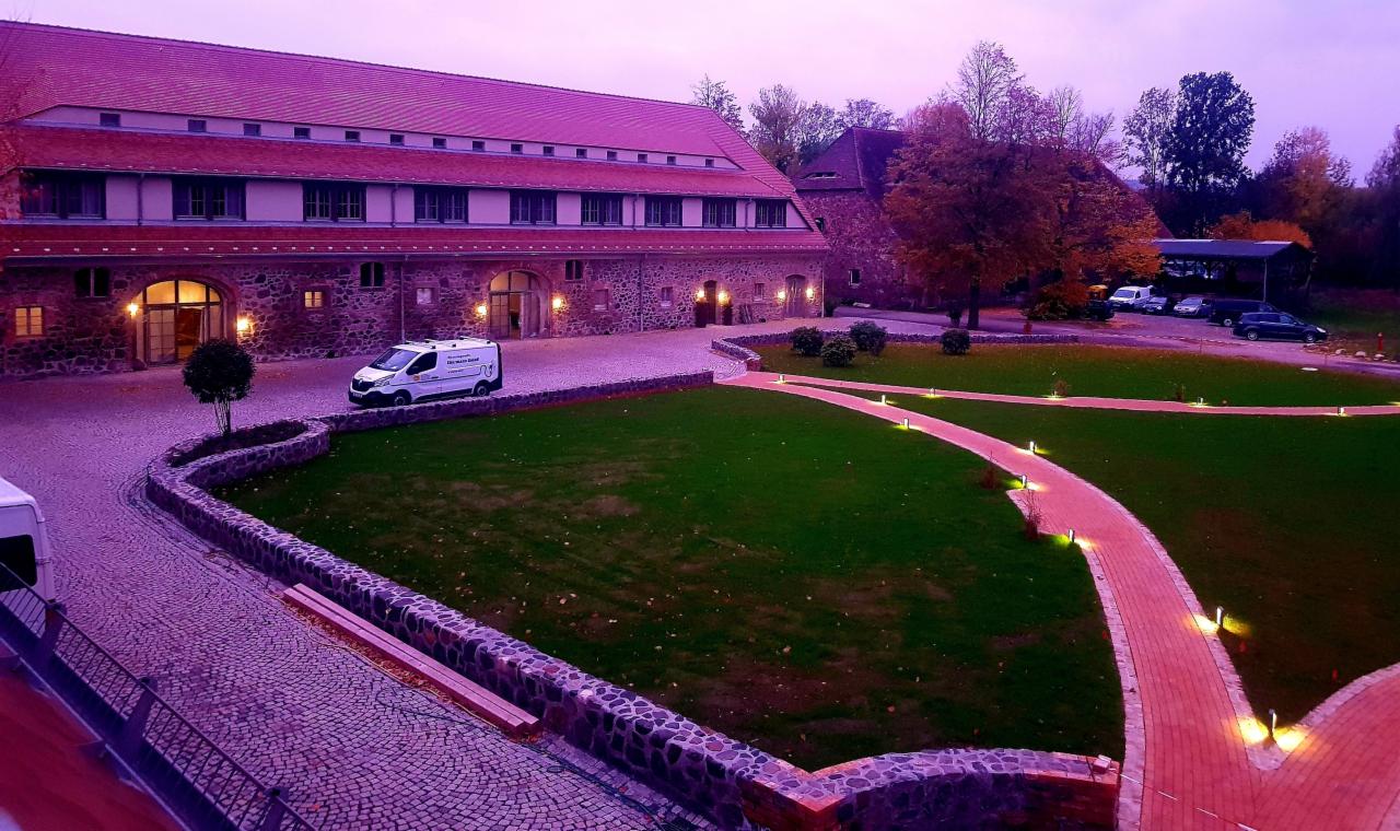 Rozległy widok na odrestaurowany, kamienny budynek z czerwonym dachem, oświetlony dziedziniec z równo przystrzyżonym trawnikiem, ścieżkami z oświetleniem punktowym i zaparkowanym samochodem...