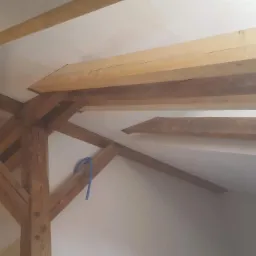 Drewniane belki stropowe w trakcie montażu na poddaszu, widoczne fragmenty białej ściany i sufitu, niebieski kabel zwisający z belki.