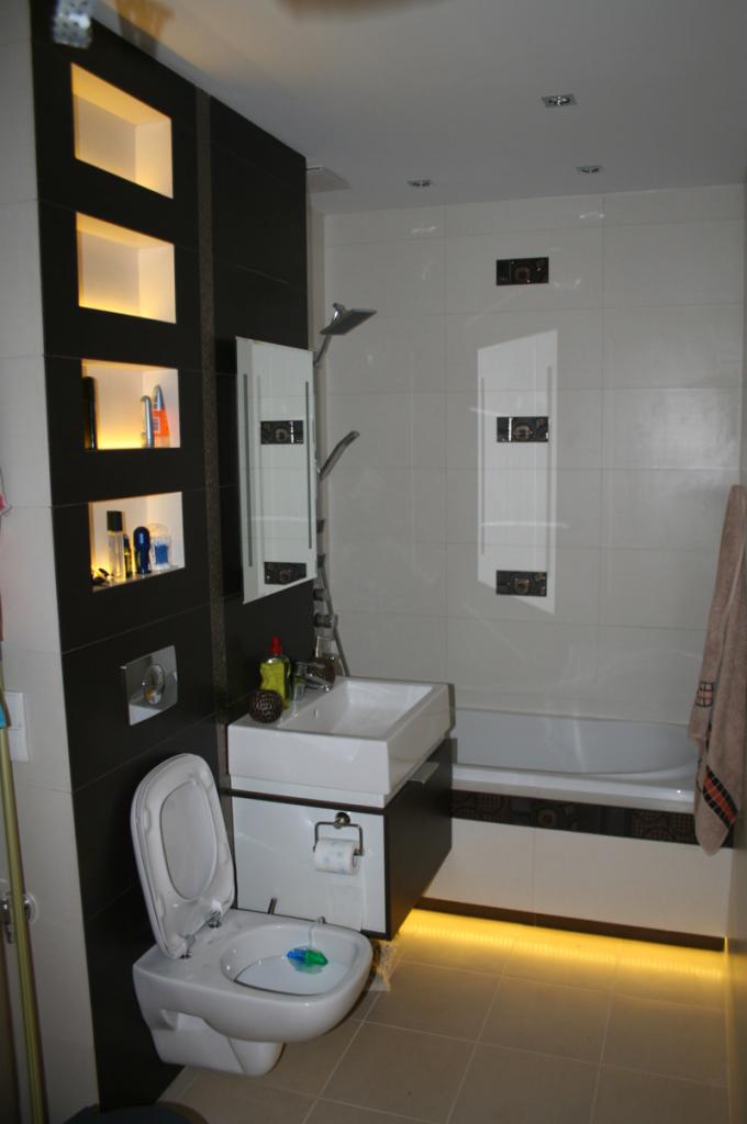 Nowoczesna łazienka z podświetlanymi półkami w czarnej zabudowie, wiszącą toaletą z niebieską kostką odświeżającą, białą umywalką nablatową i podświetlaną wanną.