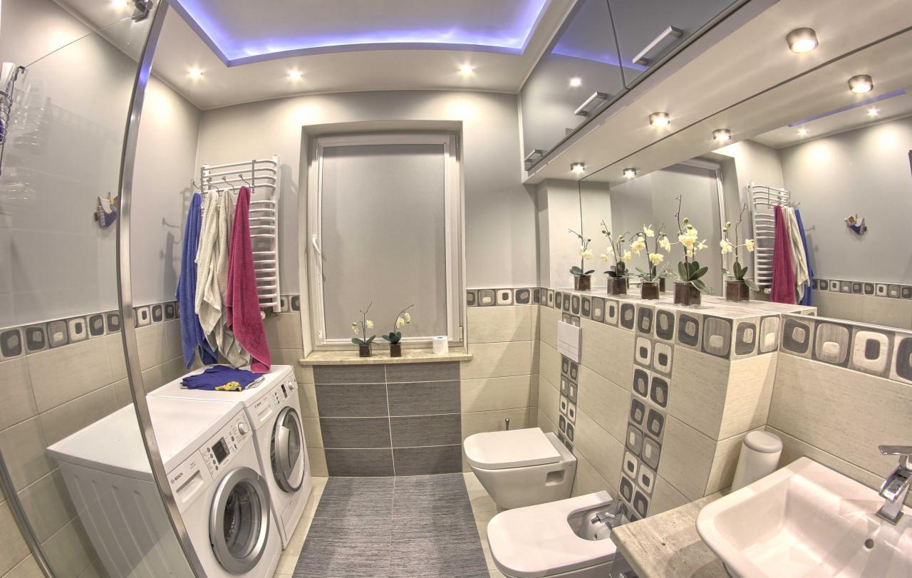 Wykończona łazienka z pralką i suszarką Bosch obok kabiny prysznicowej, szara glazura z dekoracyjnym wzorem, podświetlany sufit i kwiaty w doniczkach na parapecie.