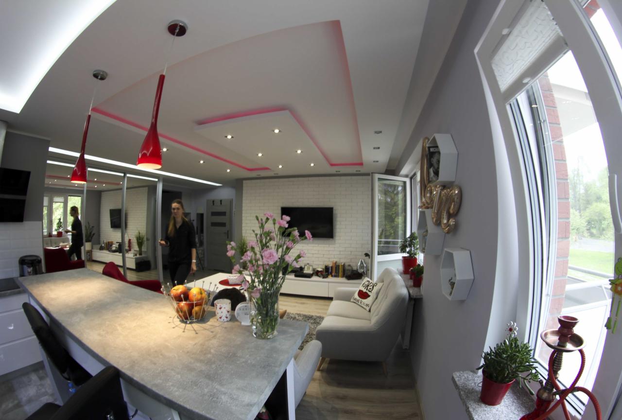 Wnętrze salonu z aneksem kuchennym, widok na wyspę kuchenną z blatem imitującym beton, czerwone lampy wiszące, dekoracyjne oświetlenie sufitu, biała cegła na ścianie, kobieta w tle, szeroki kąt...