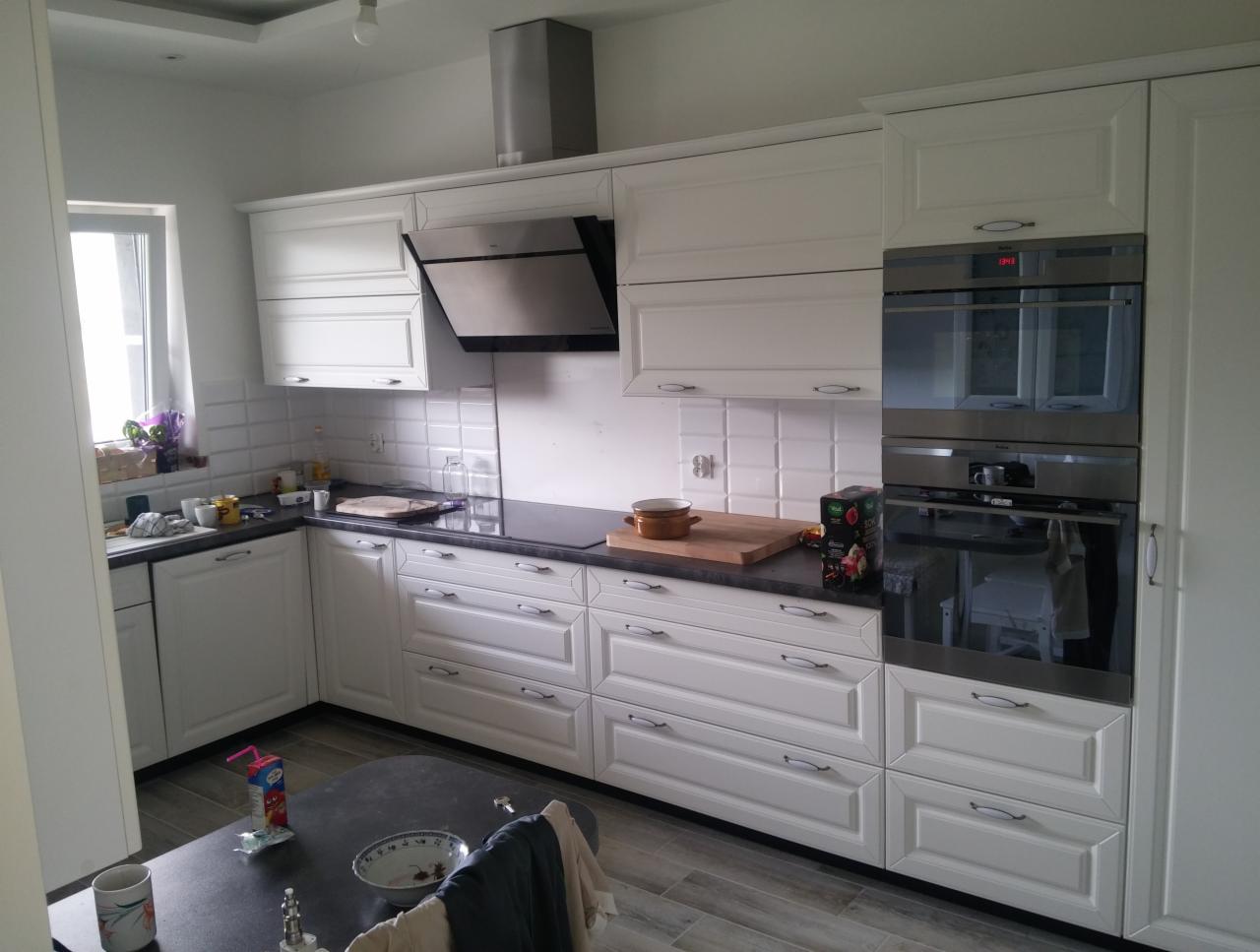 Jasna kuchnia z białymi szafkami w stylu klasycznym, czarnym blatem i płytkami na ścianie, widoczne dwa piekarniki w zabudowie i okap nad płytą grzewczą.