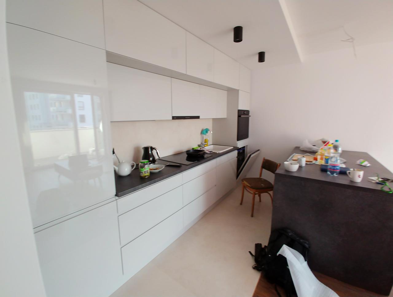 Nowoczesna biała kuchnia z czarnym blatem, widoczne czajniki, zlewozmywak i wyspa kuchenna z drobnymi przedmiotami, w tle krzesło i niedokończone wykończenie sufitu.