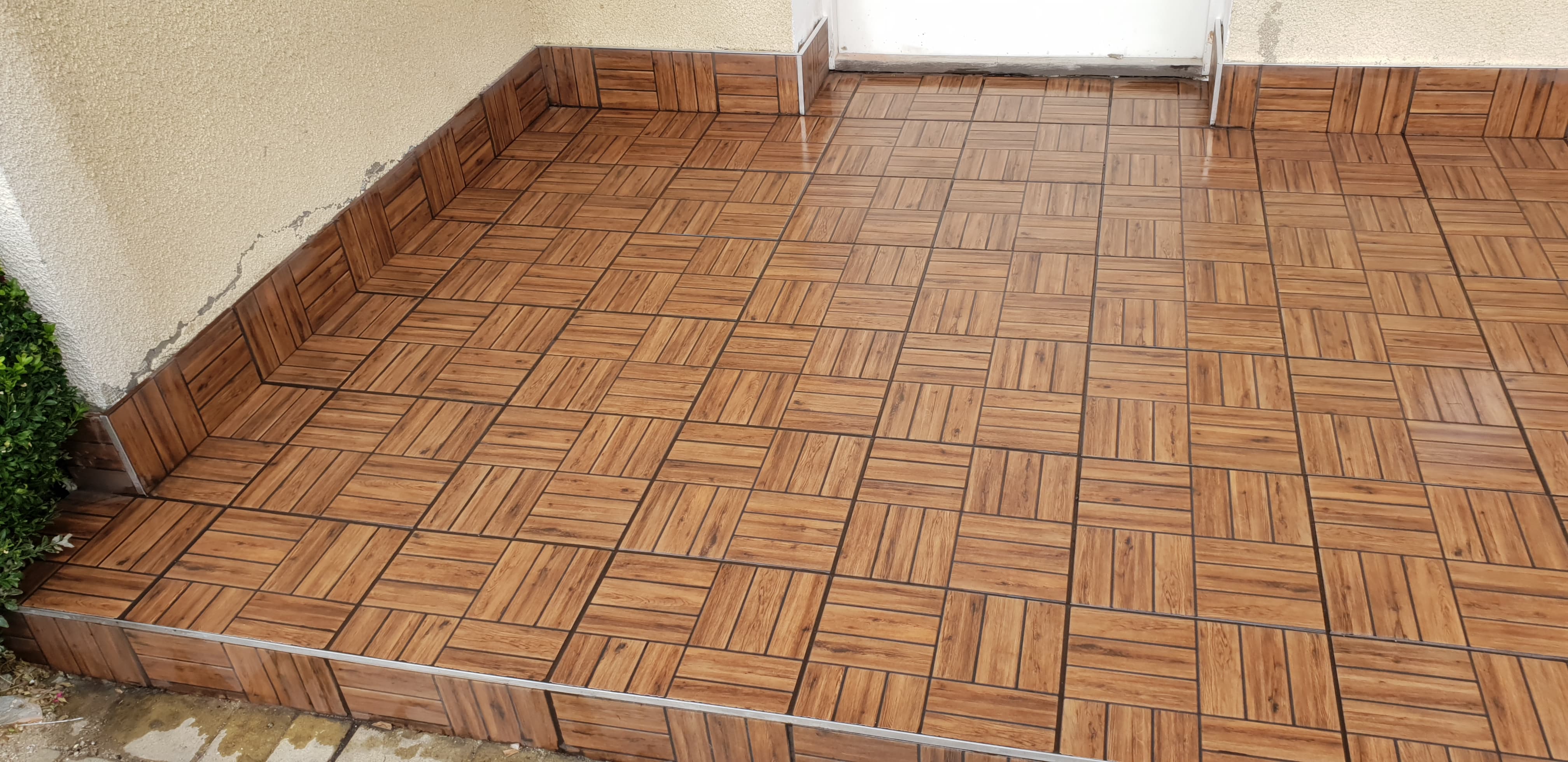 Ułożona pod kątem brązowa terakota imitująca drewno na zewnątrz budynku, z widocznym fragmentem ściany i zieleni po lewej stronie oraz białymi drzwiami w tle.