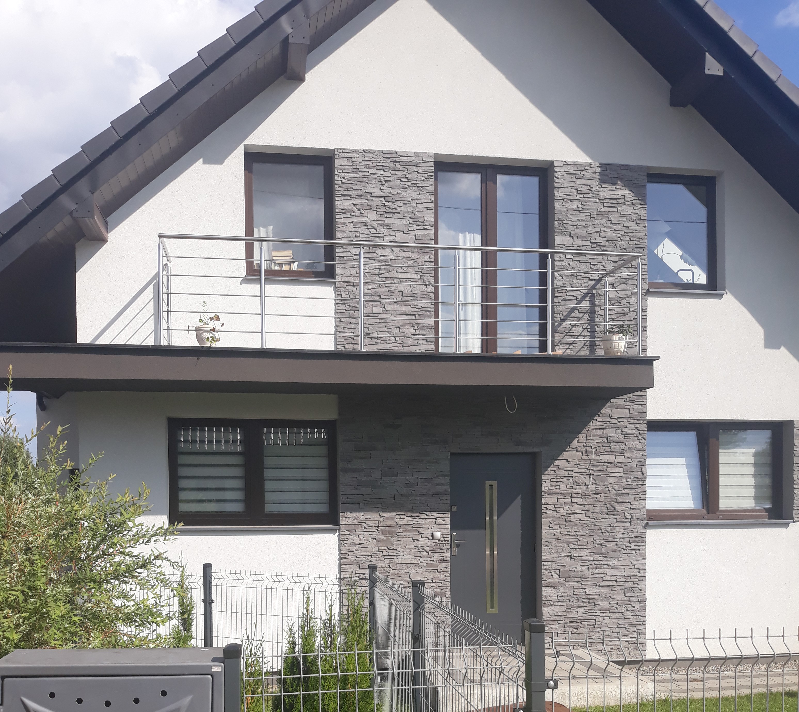 Dwupiętrowy dom jednorodzinny z białą elewacją i szarą okładziną imitującą kamień, balkon z metalową balustradą, szara stolarka okienna i drzwiowa, ogrodzenie metalowe.
