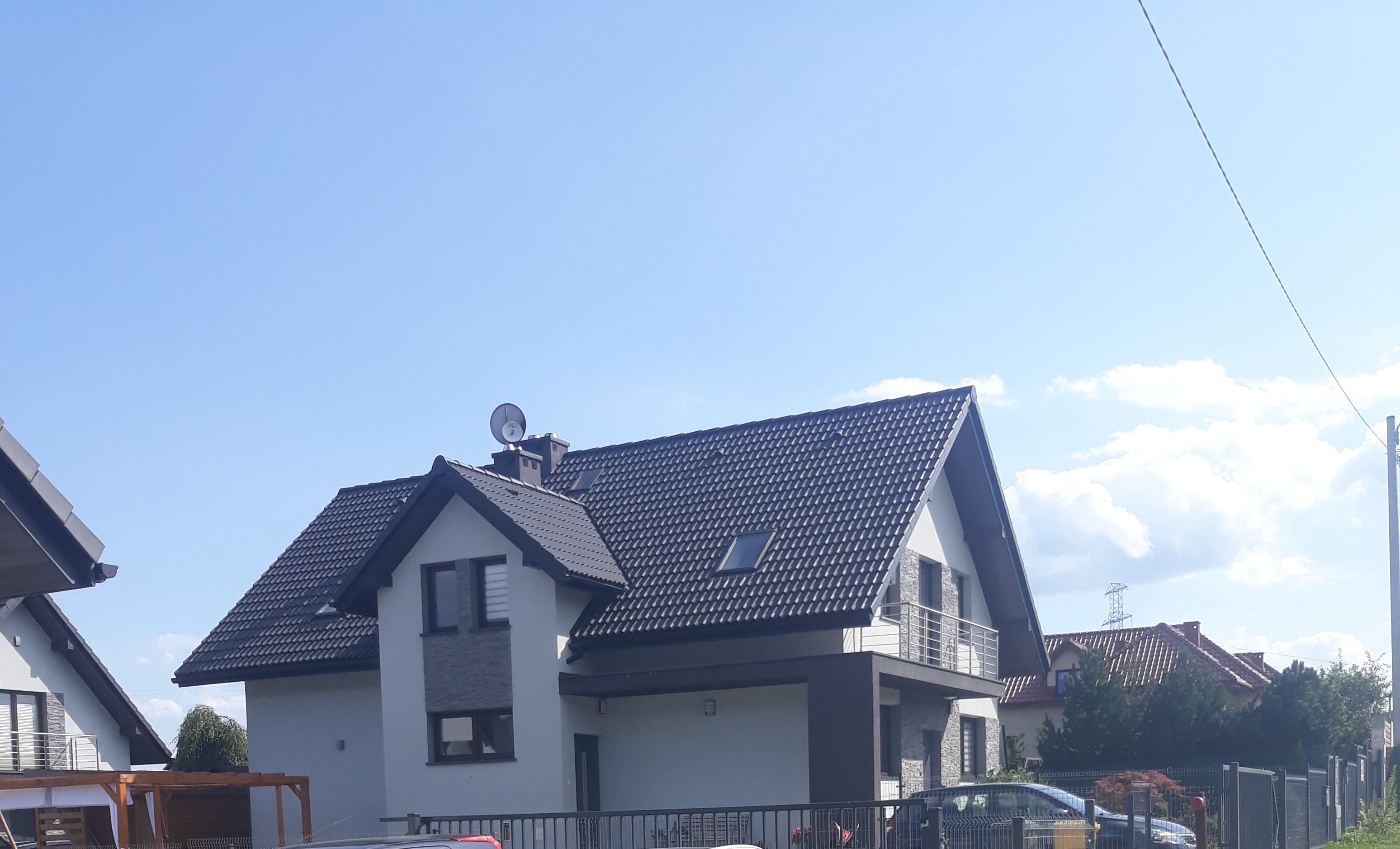 Nowoczesny dom jednorodzinny z grafitową dachówką ceramiczną, jasną elewacją i balkonem z metalową balustradą, widoczny zza ogrodzenia.