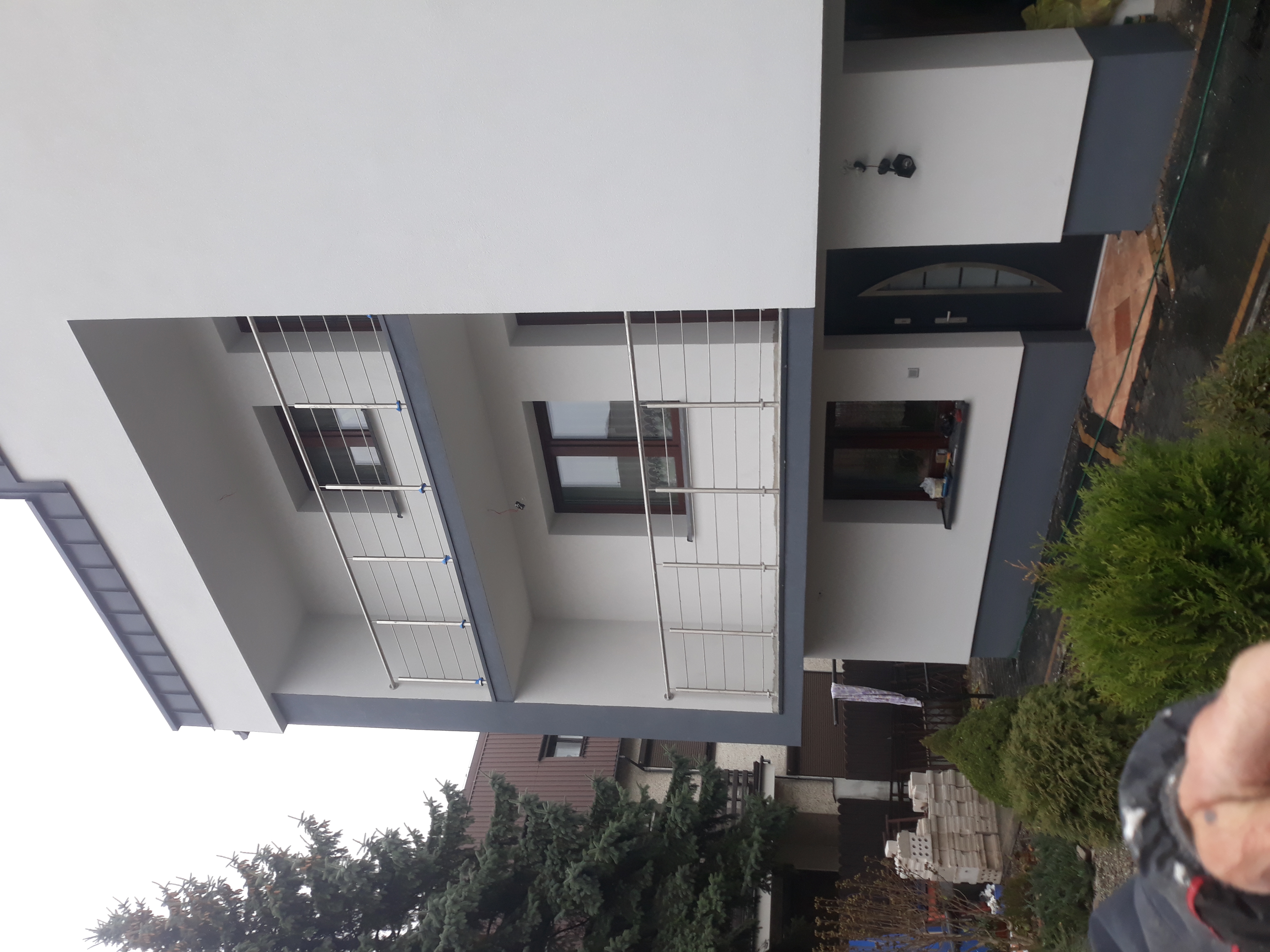 Nowoczesny dom z jasną elewacją, balkonami z metalowymi balustradami i ciemnoszarą stolarką okienną i drzwiową.