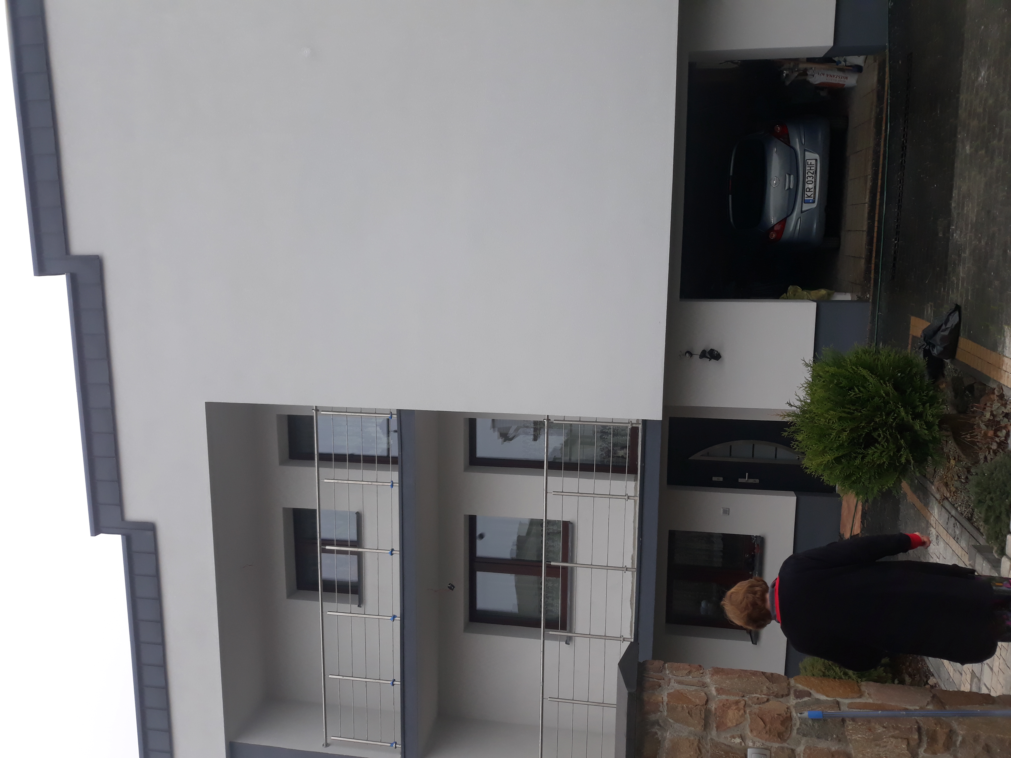 Dwupiętrowy dom z garażem i balkonami z metalowymi barierkami, świeżo otynkowany na biało, z szarą podbitką dachową, z widocznym fragmentem kamiennego ogrodzenia i osoby w czarnym płaszczu.