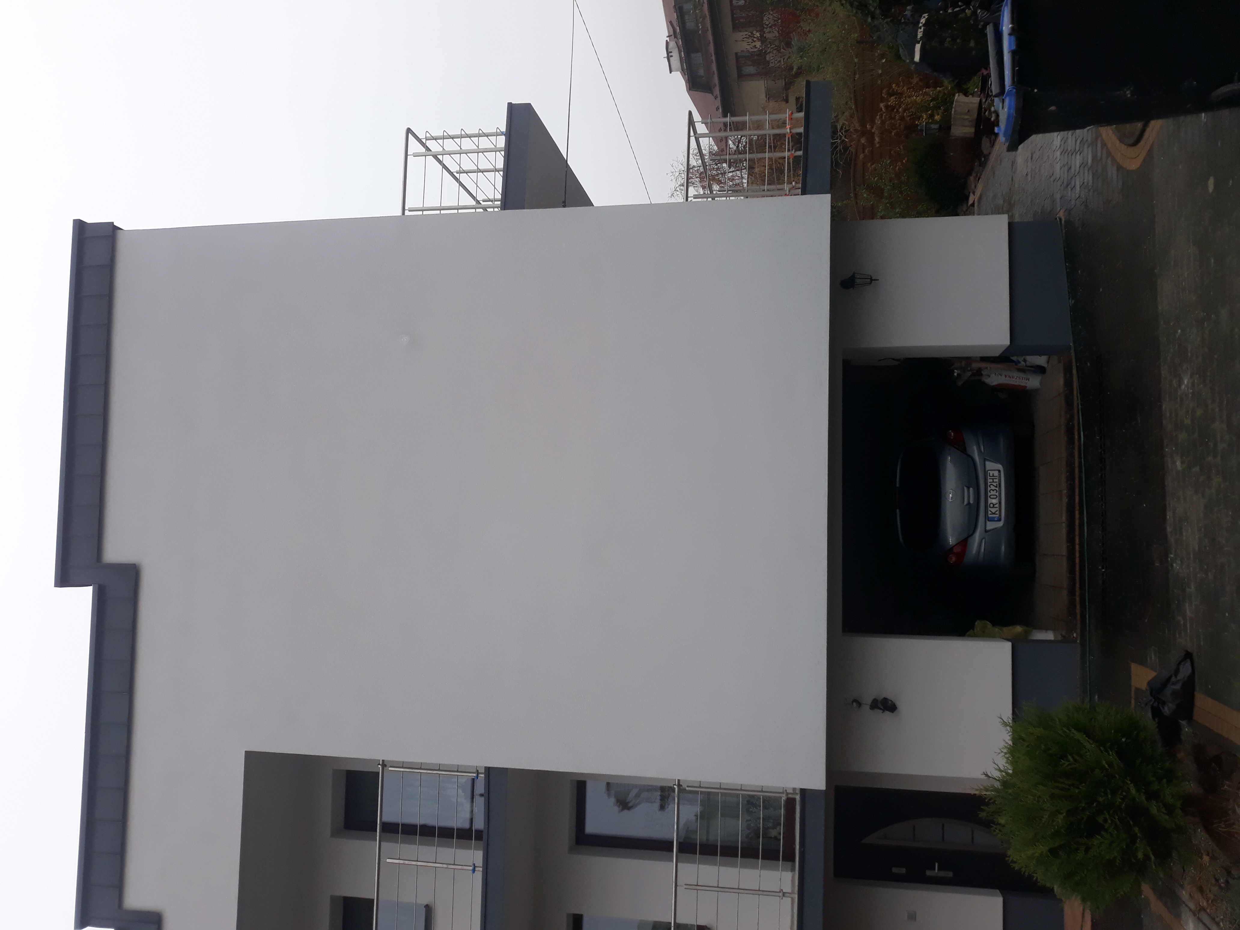 Nowoczesny dom z białą elewacją i szarymi akcentami, widok z zewnątrz, samochód w garażu, widoczne okna z balustradami.