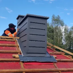 Dekarz montuje obróbkę blacharską komina na dachu z częściowo ułożoną dachówką ceramiczną w kolorze grafitowym, widoczna więźba dachowa i czerwona membrana dachowa, na tle błękitne niebo z chmurami...