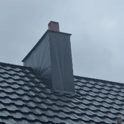 Nowy, ciemnoszary dach z blachy trapezowej i obróbka komina z blachy w geometrycznym stylu, widoczne nity mocujące, pochmurne niebo w tle.