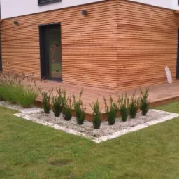 Nowoczesny dom z elewacją z jasnego drewna, tarasem i starannie zagospodarowanym ogrodem z niskimi krzewami iglastymi na białym żwirze i ozdobnymi trawami.