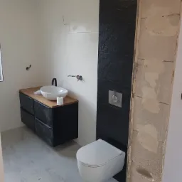 Wnętrze łazienki w trakcie remontu: białe płytki, czarna ściana z teksturą, podwieszana toaleta, umywalka nablatowa na drewnianym blacie z czarną szafką, widoczne niedokończone prace na ścianie.