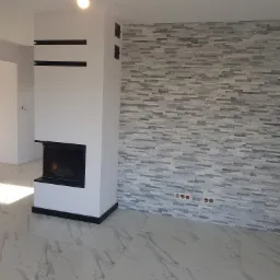 Nowoczesny salon z białym kominkiem z czarnymi akcentami, ściana wyłożona szarymi płytkami imitującymi kamień, marmurowa podłoga, surowe oświetlenie.