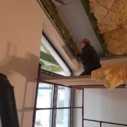 Pracownik na rusztowaniu montuje izolację termiczną poddasza przy oknie dachowym, widoczna wełna mineralna i folia paroizolacyjna.