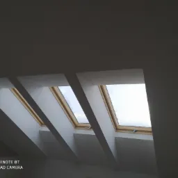 Trzy okna dachowe z drewnianymi ramami w białej zabudowie kartonowo-gipsowej, widok od dołu pod kątem, lekko szare tło ścian.