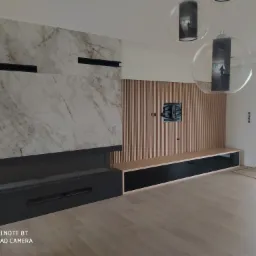 Nowoczesny salon w Wąsoszu Górnym z marmurową ścianą kominkową, drewnianymi lamelami na ścianie TV i minimalistyczną szafką RTV z czarnymi frontami, uzupełniony o wiszące lampy kuliste.