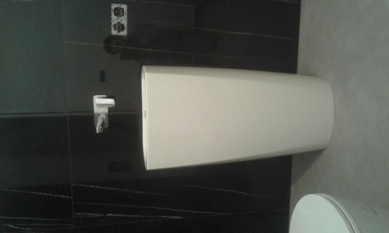 Biała, stojąca umywalka w łazience z czarnymi, błyszczącymi płytkami na ścianie i jasnoszarą podłogą. Widoczny kran i gniazdka elektryczne.