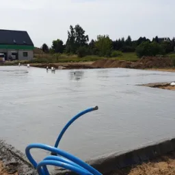 Świeżo wylana, mokra płyta fundamentowa domu jednorodzinnego na działce z widocznymi elementami szalunku i niebieskim wężem do pompy betonu.