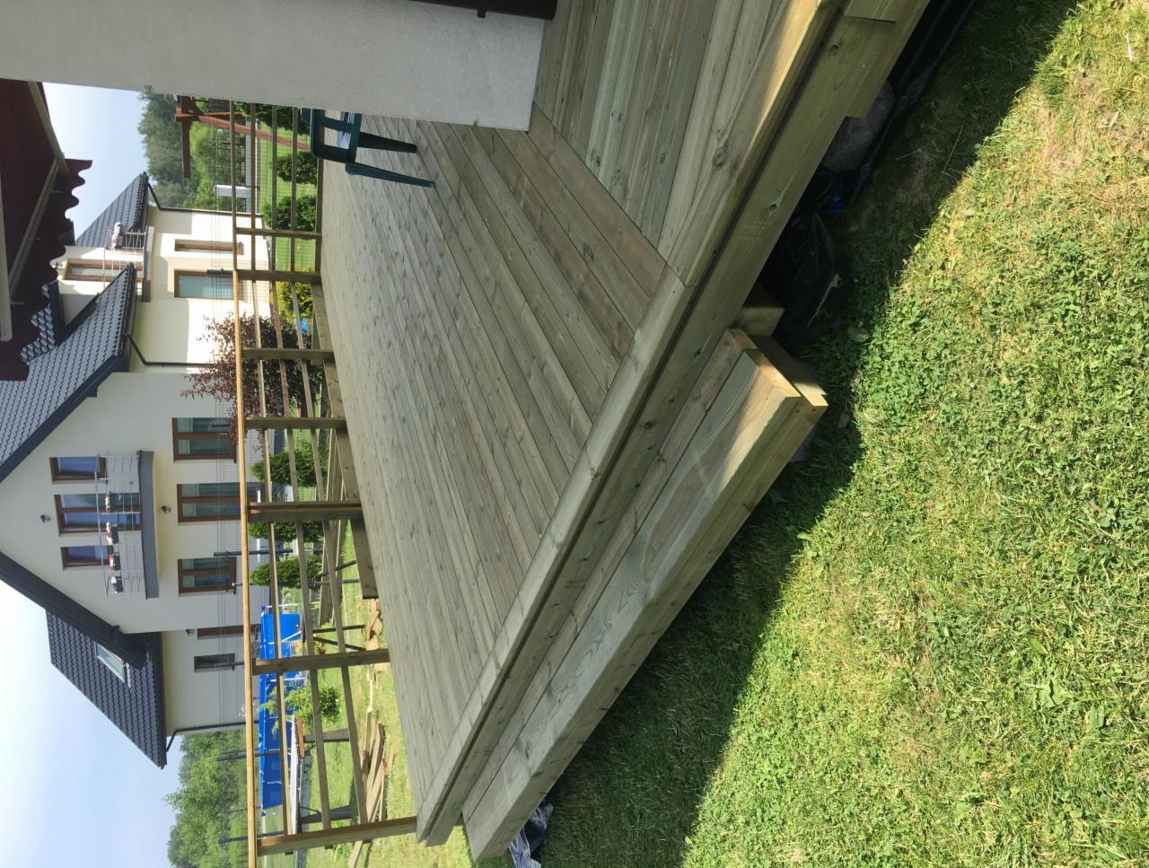 Zewnętrzny taras drewniany z desek ułożonych pod kątem, widok z poziomu trawnika, w tle dom jednorodzinny z balkonem i zielony ogród.