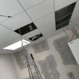 Montaż sufitu podwieszanego z płyt karton-gips w biurze, widoczne otwory na wentylację i oświetlenie, ściany w trakcie szpachlowania, drabina oparta o ścianę.