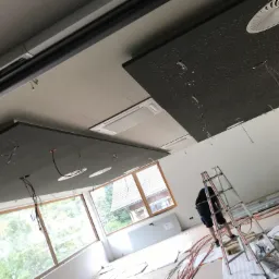 Montaż sufitu podwieszanego z panelami akustycznymi i instalacją elektryczną, widoczne kable i drabina w pomieszczeniu z oknami.