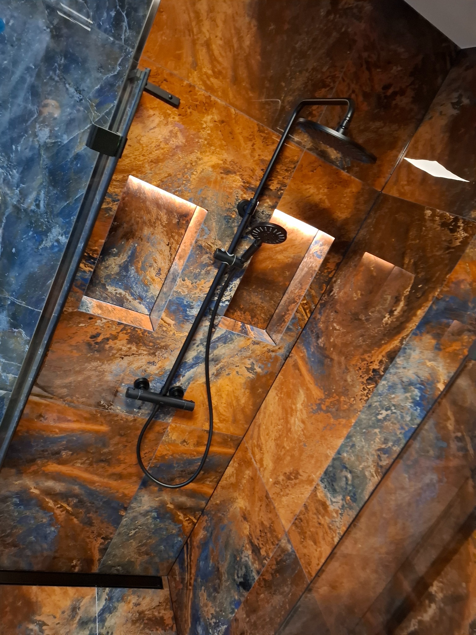 Nowoczesna kabina prysznicowa z oryginalnymi płytkami w odcieniach brązu i niebieskiego, z wnękami podświetlonymi ledami i czarną armaturą.