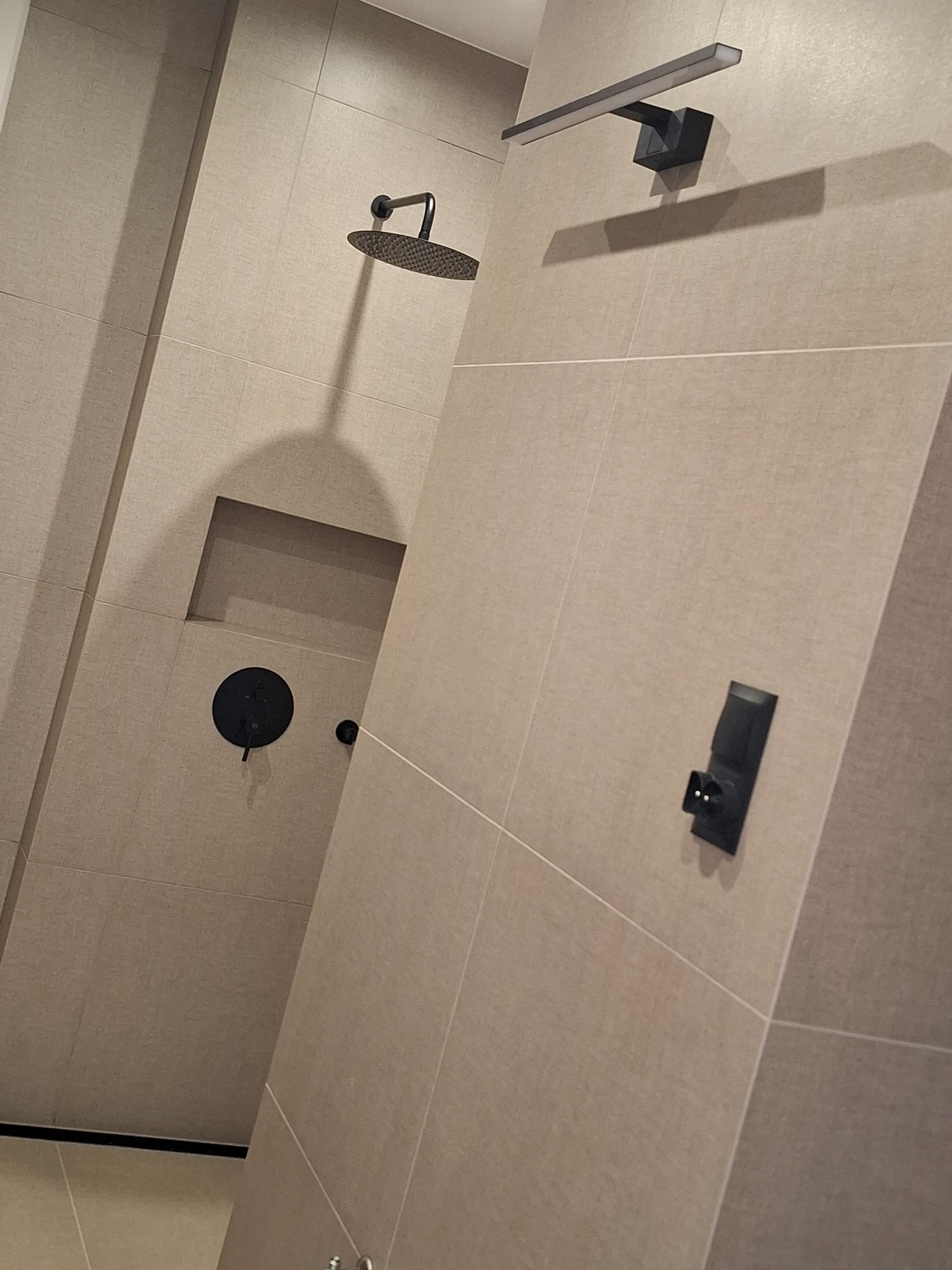 Beżowa, nowoczesna łazienka z czarną armaturą: deszczownica, bateria, oświetlenie LED i gniazdko. Ułożone płytki tworzą minimalistyczny design wnętrza.