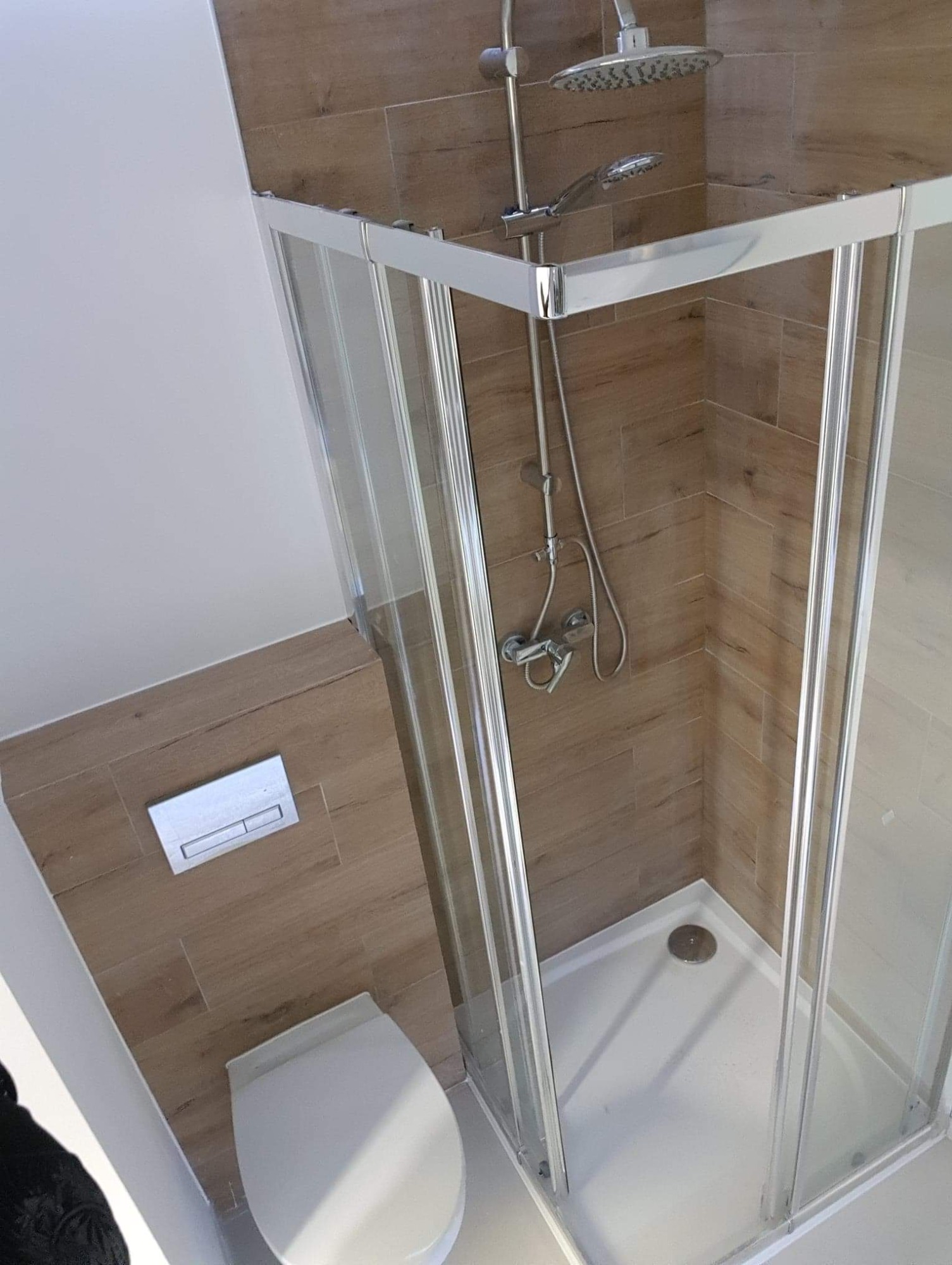 Nowoczesna kabina prysznicowa z transparentnymi drzwiami, wykończona płytkami imitującymi drewno, z widoczną toaletą w tle, minimalistyczny design.