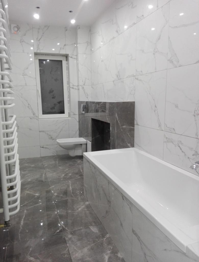 Nowoczesna łazienka wykończona płytkami imitującymi marmur, z grzejnikiem drabinkowym, zabudową wnęki nad toaletą i wanną.