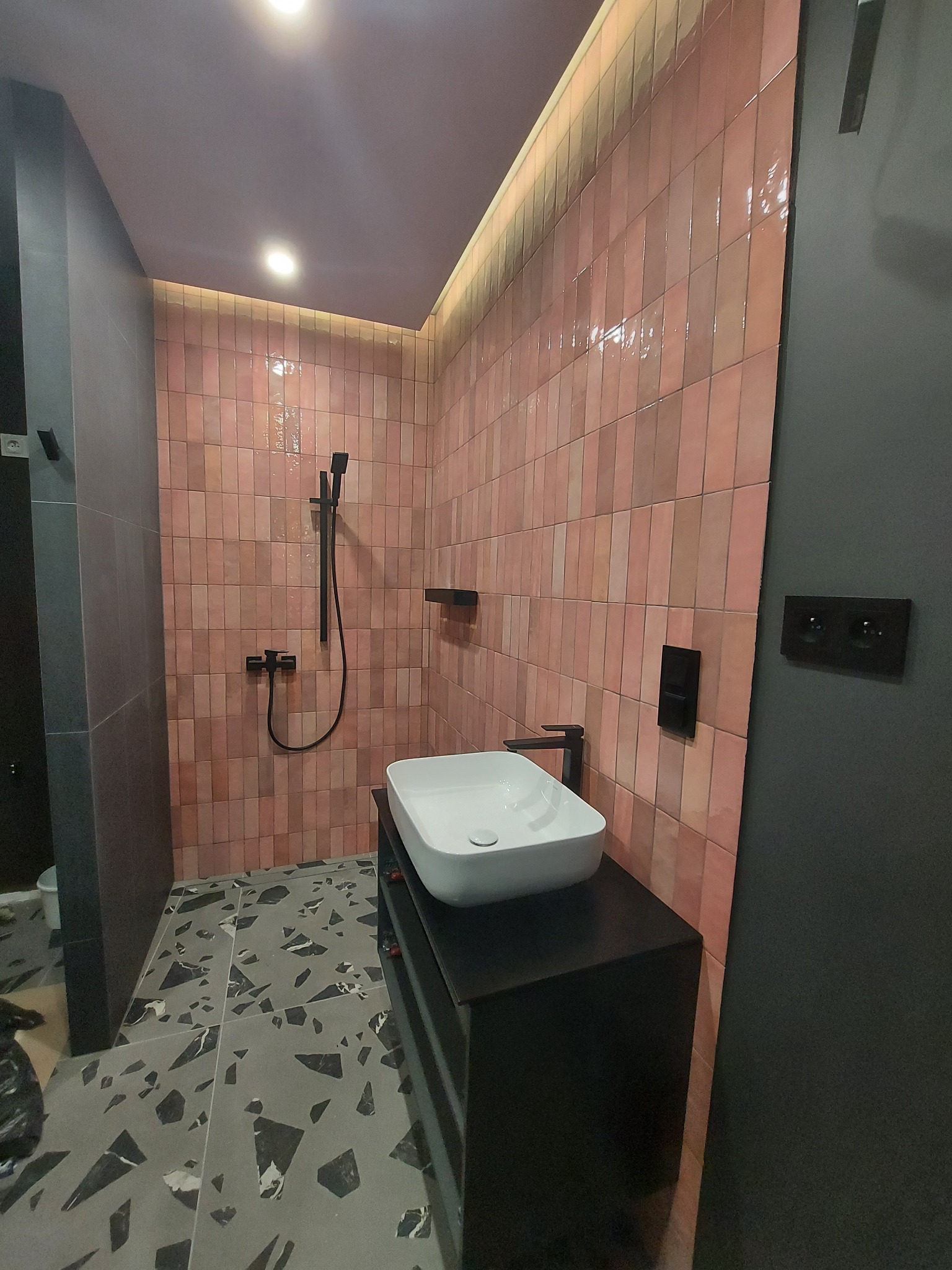 Nowoczesna łazienka z różowymi, pionowo ułożonymi płytkami, czarną armaturą i szarą posadzką z motywem lastryko, oświetlona listwą LED pod sufitem.