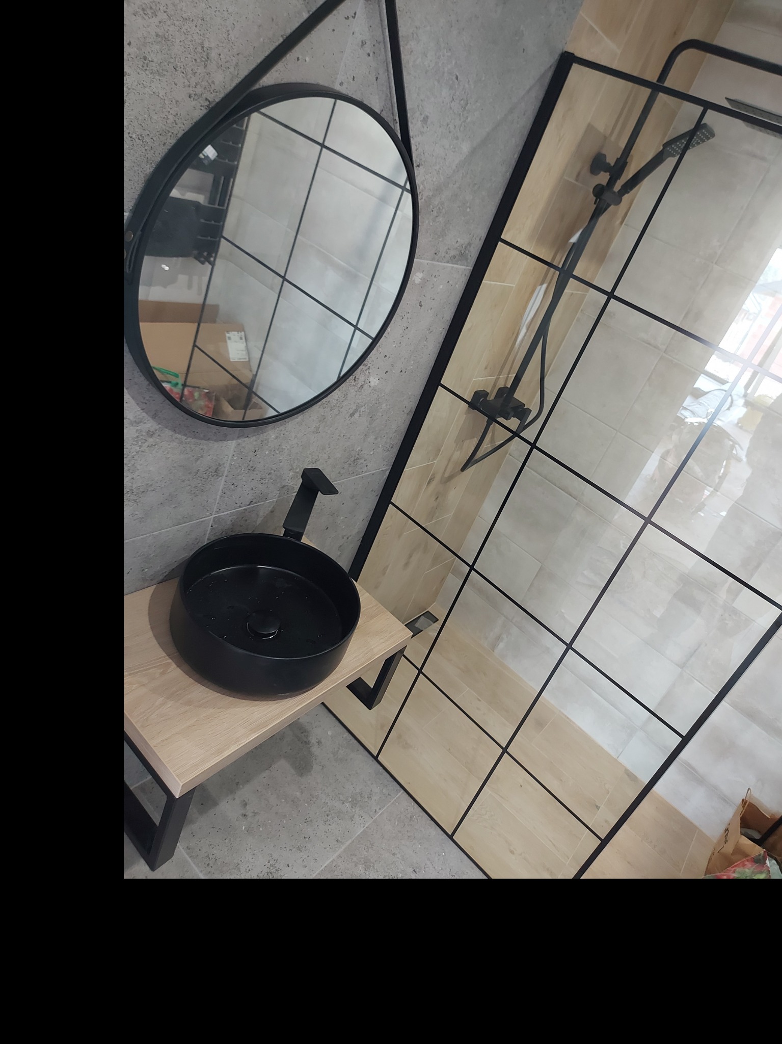 Nowoczesna łazienka z czarną umywalką nablatową, okrągłym lustrem w czarnej ramie i kabiną prysznicową typu walk-in z czarnymi profilami.