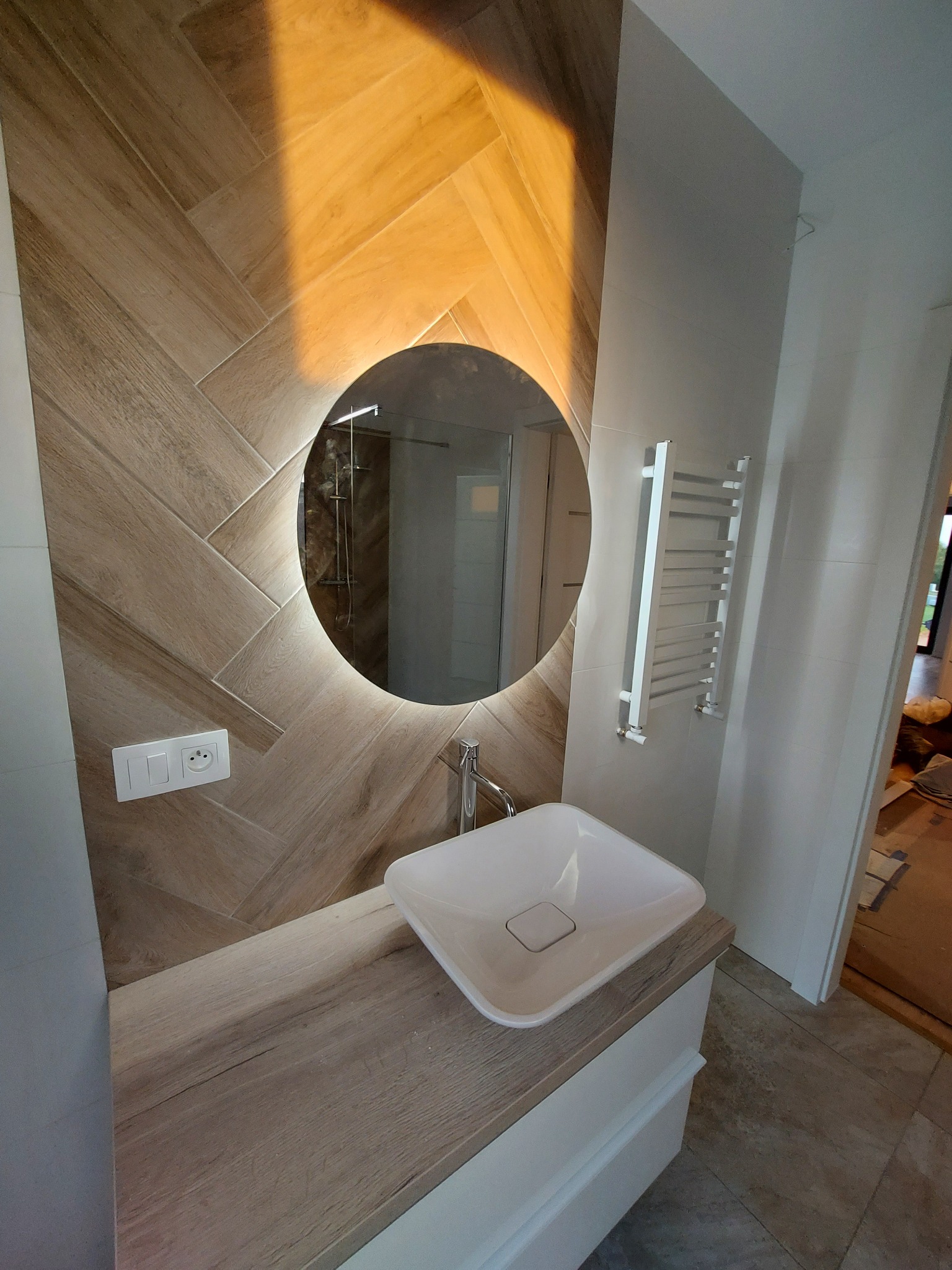 Nowoczesna łazienka z umywalką nablatową, podświetlanym okrągłym lustrem i drewnopodobnymi płytkami ułożonymi w jodełkę na ścianie.