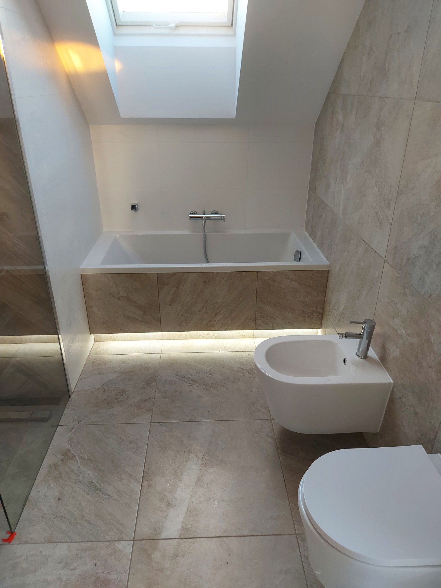 Nowoczesna łazienka z oknem dachowym, wykończona płytkami imitującymi marmur, z podświetloną wanną i białym bidetem.