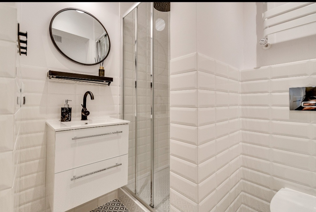 Biała łazienka z kabiną prysznicową, umywalką z szafką, czarną armaturą i geometrycznym wzorem na podłodze.