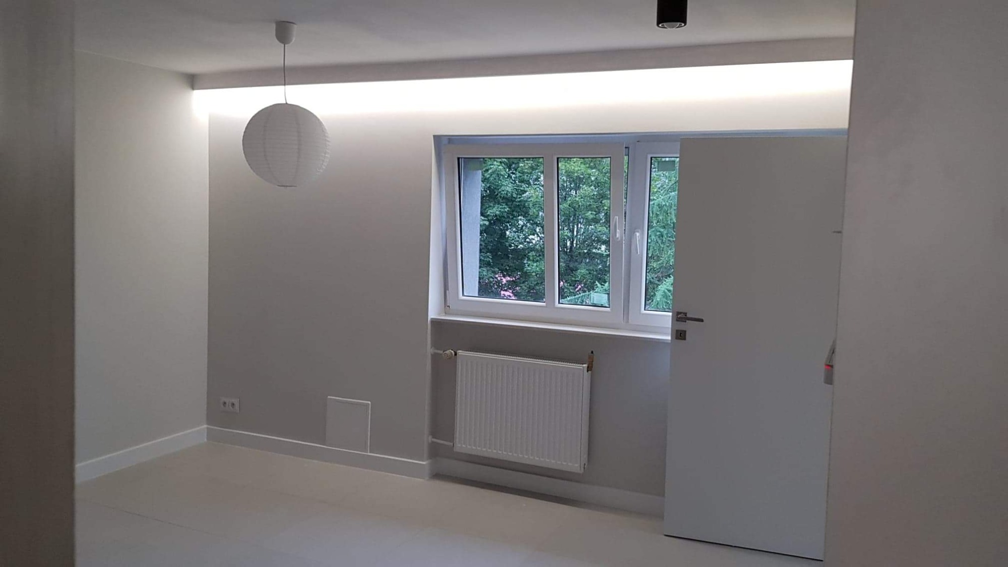 Jasne, odświeżone wnętrze pokoju z białymi ścianami, podświetlanym sufitem i lampionem, widok na okno z zielenią na zewnątrz.