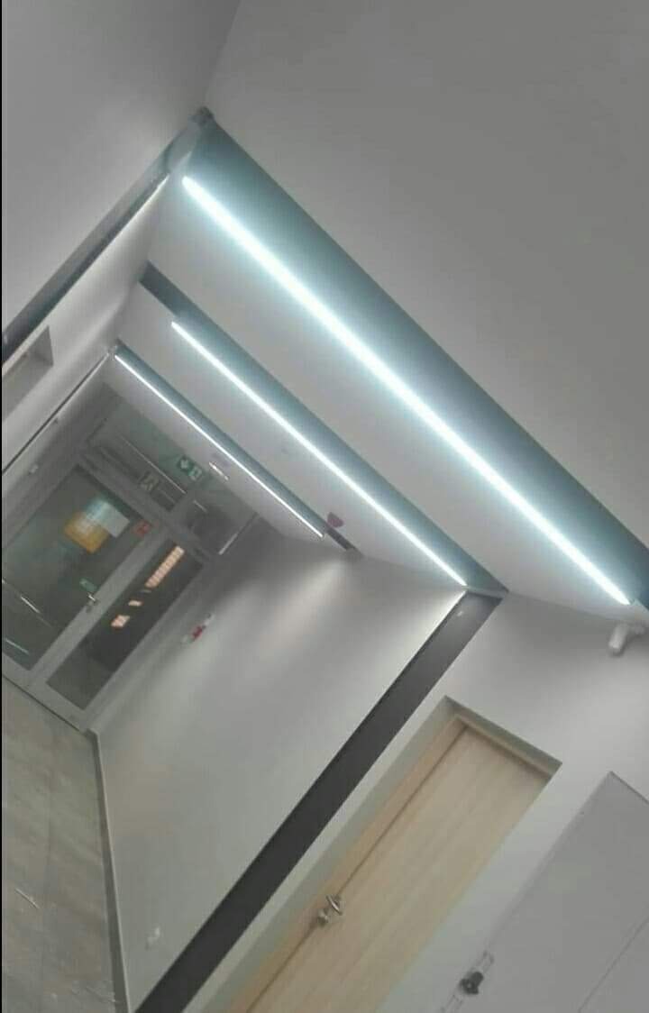 Korytarz biurowy z zamontowanym oświetleniem LED liniowym wzdłuż sufitu i ściany, widoczna drewniana ościeżnica drzwiowa i przeszklone wejście do pomieszczenia.