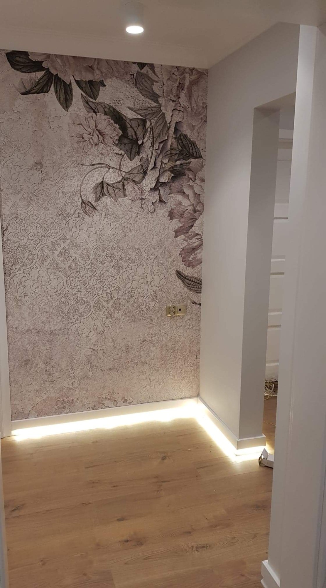 Ściana z dekoracyjną tapetą w kwiaty, oświetlenie LED przy podłodze, widok na korytarz z białymi ścianami.