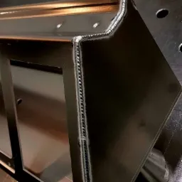 Fragment spawanej konstrukcji z metalu, widoczne precyzyjne spoiny na krawędziach, całość umieszczona na perforowanym stole spawalniczym.