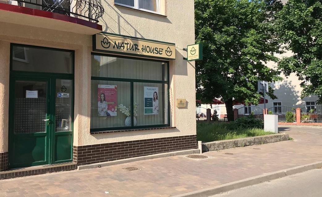 Fasada budynku Natur House z zielonymi drzwiami i oknami, szyld z logo i plakaty w oknach.