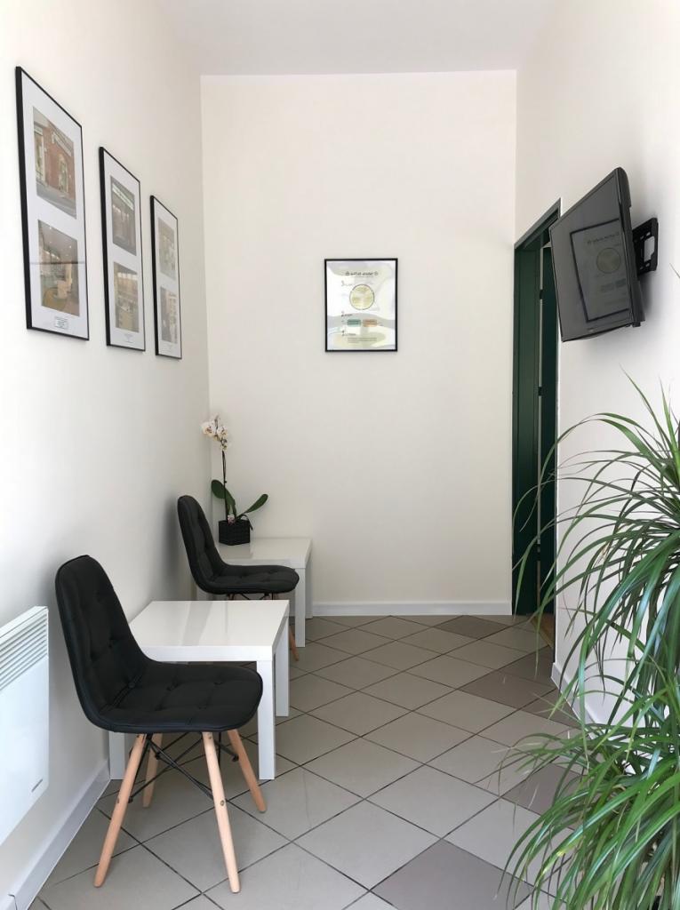 Poczekalnia w gabinecie: Dwa czarne krzesła z drewnianymi nogami przy białych stolikach, na ścianach grafiki w czarnych ramkach, na podłodze płytki w szaro-białą kratę, roślina doniczkowa w rogu.