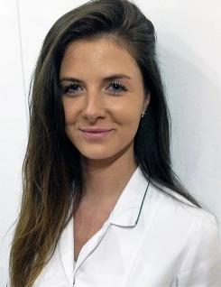 Portret młodej kobiety w białym uniformie z ciemnymi włosami, uśmiechająca się, na jasnym tle.